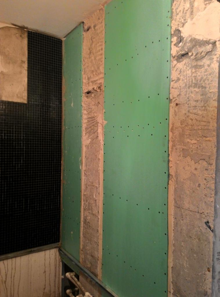 Montaż zielonych płyt kartonowo-gipsowych na ścianie łazienki, widoczne fragmenty starej instalacji i czarne płytki mozaikowe.