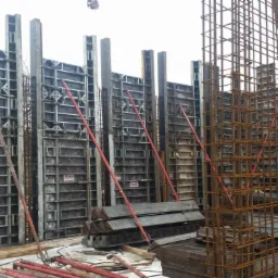 Szare szalunki pionowe z metalowymi wzmocnieniami i czerwonymi podporami, otoczone siatką zbrojeniową na placu budowy w zimowy dzień.