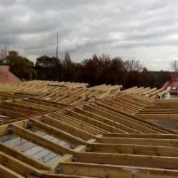 Konstrukcja drewniana dachu w trakcie budowy, widoczne krokwie, murłaty i bloczki betonowe, w tle zabudowa miejska i żuraw budowlany pod pochmurnym niebem.