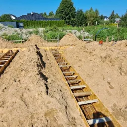 Przygotowanie fundamentów pod budowę domu: szalunki zbrojeniowe wypełnione ziemią, widoczne instalacje wodne, w tle zieleń i zabudowania.