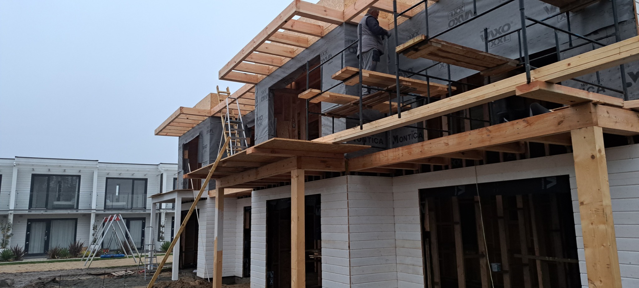 Wznoszenie dwukondygnacyjnego domu w zabudowie szeregowej z drewnianą konstrukcją szkieletową, widoczne rusztowania i pracownik w trakcie montażu. W tle gotowe segmenty z balkonami.