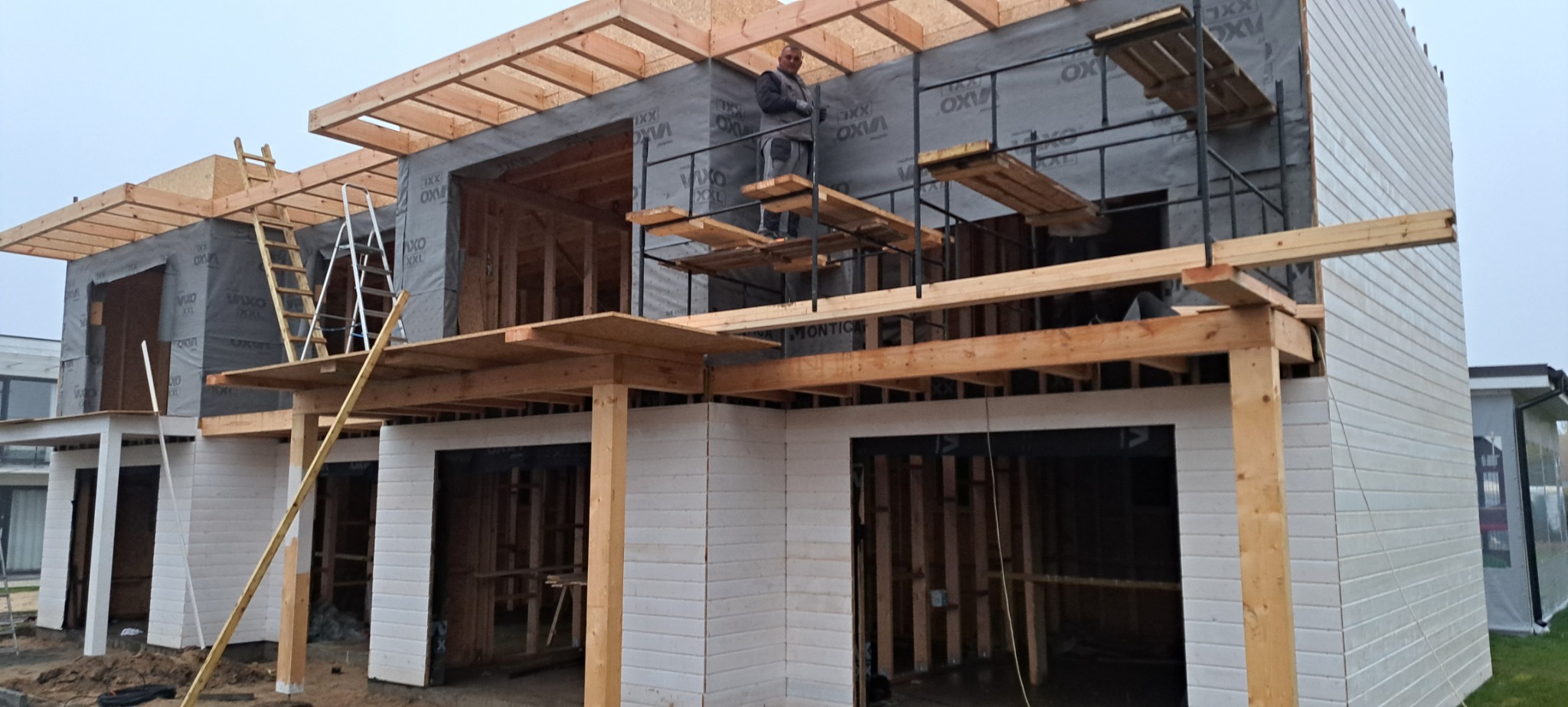 Budowa domu w stanie surowym otwartym: widoczne drewniane elementy konstrukcyjne, ocieplenie ścian folią, rusztowania i pracownik na wysokości