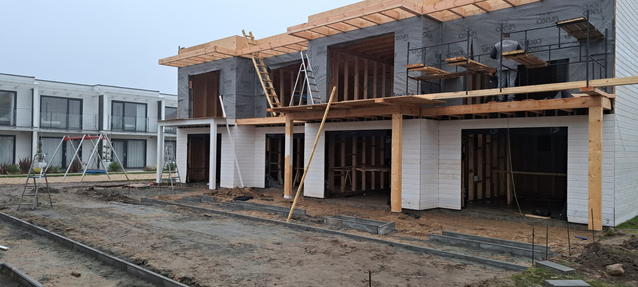 Budowa segmentów domów w trakcie realizacji: widoczne drewniane konstrukcje, folia ochronna na ścianach, rusztowania i praca na wysokościach, zarys osiedla w tle.