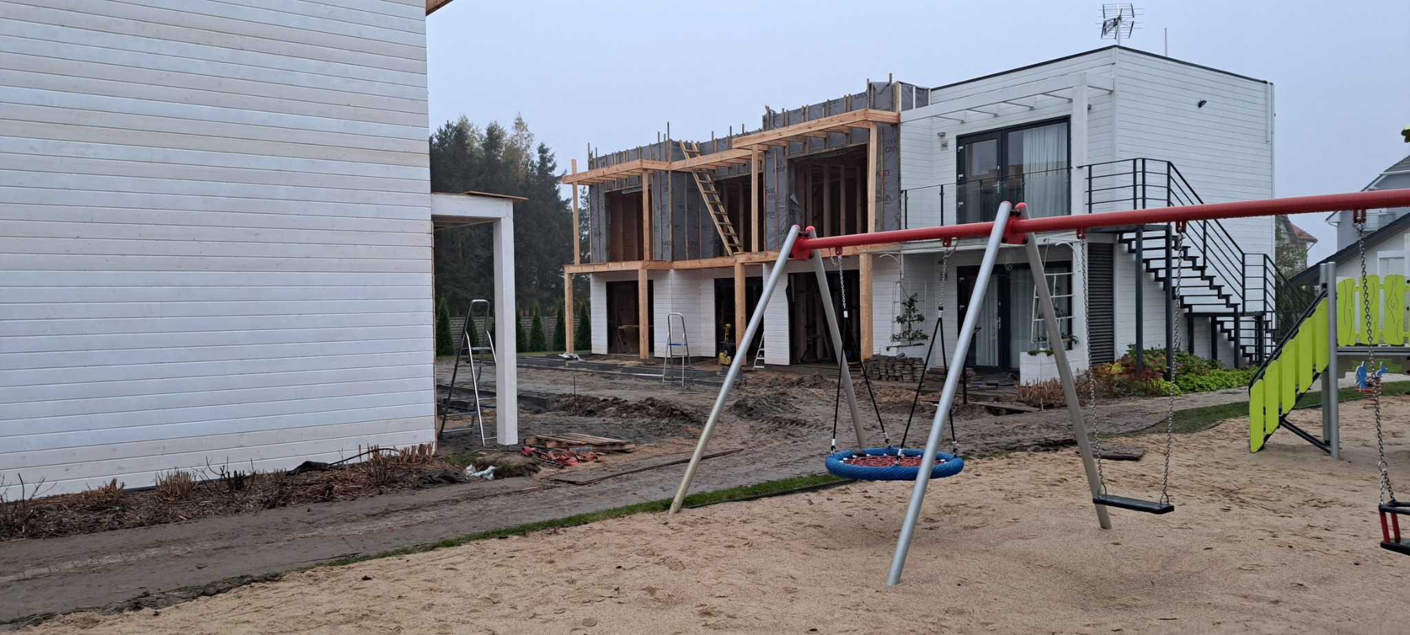 Stan surowy zamknięty bliźniaka w Koszalinie, widok z placu zabaw, konstrukcja drewniana dachu, biała elewacja z drewnianą boazerią, schody zewnętrzne.