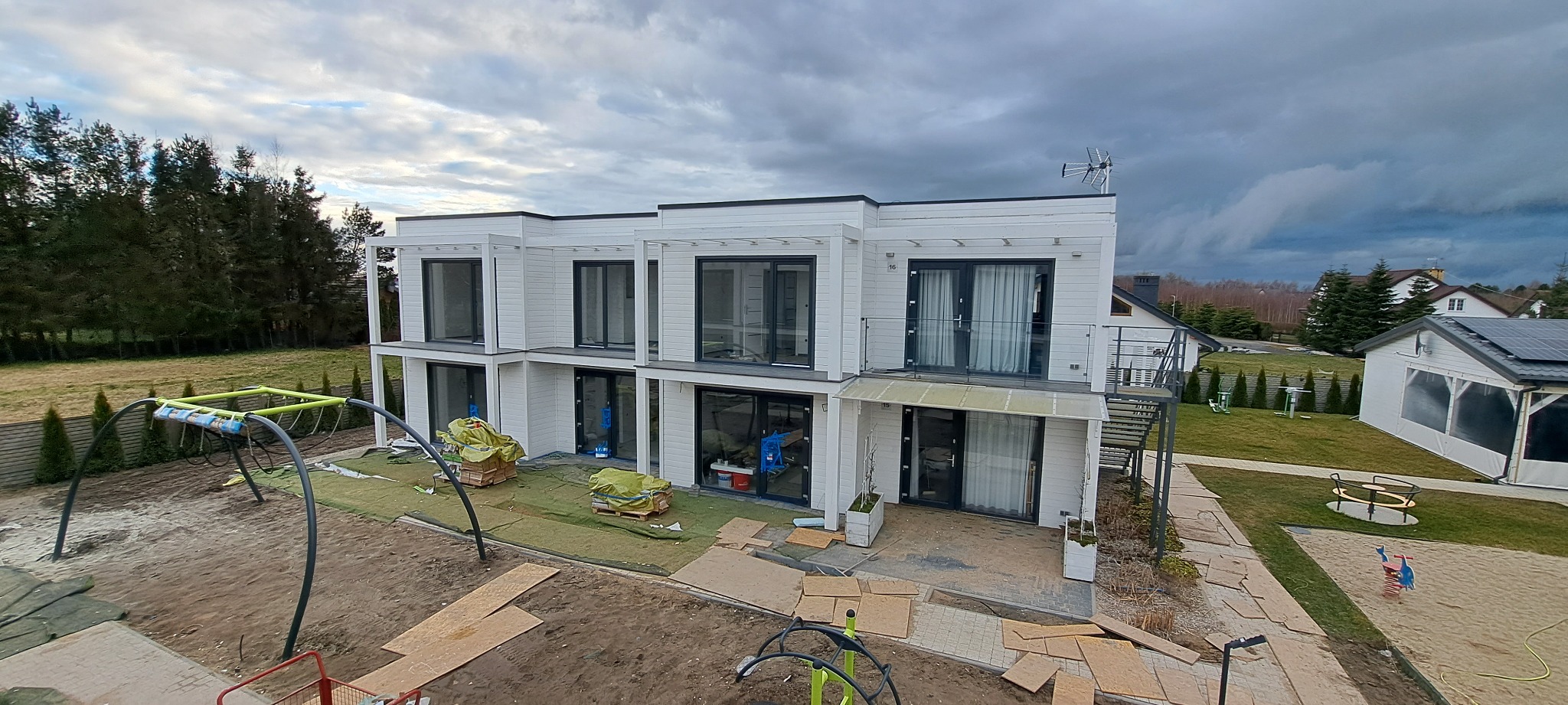 Nowoczesny, dwukondygnacyjny dom w zabudowie szeregowej z białymi elewacjami i dużymi oknami, w trakcie prac wykończeniowych na zewnątrz, widoczny plac zabaw w tle i pochmurne niebo.