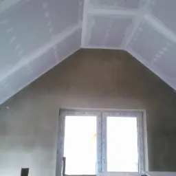 Poddasze z oknem, ściany pokryte szpachlą, sufit w trakcie montażu płyt gipsowo-kartonowych, widoczne łączenia płyt wymagające wykończenia.