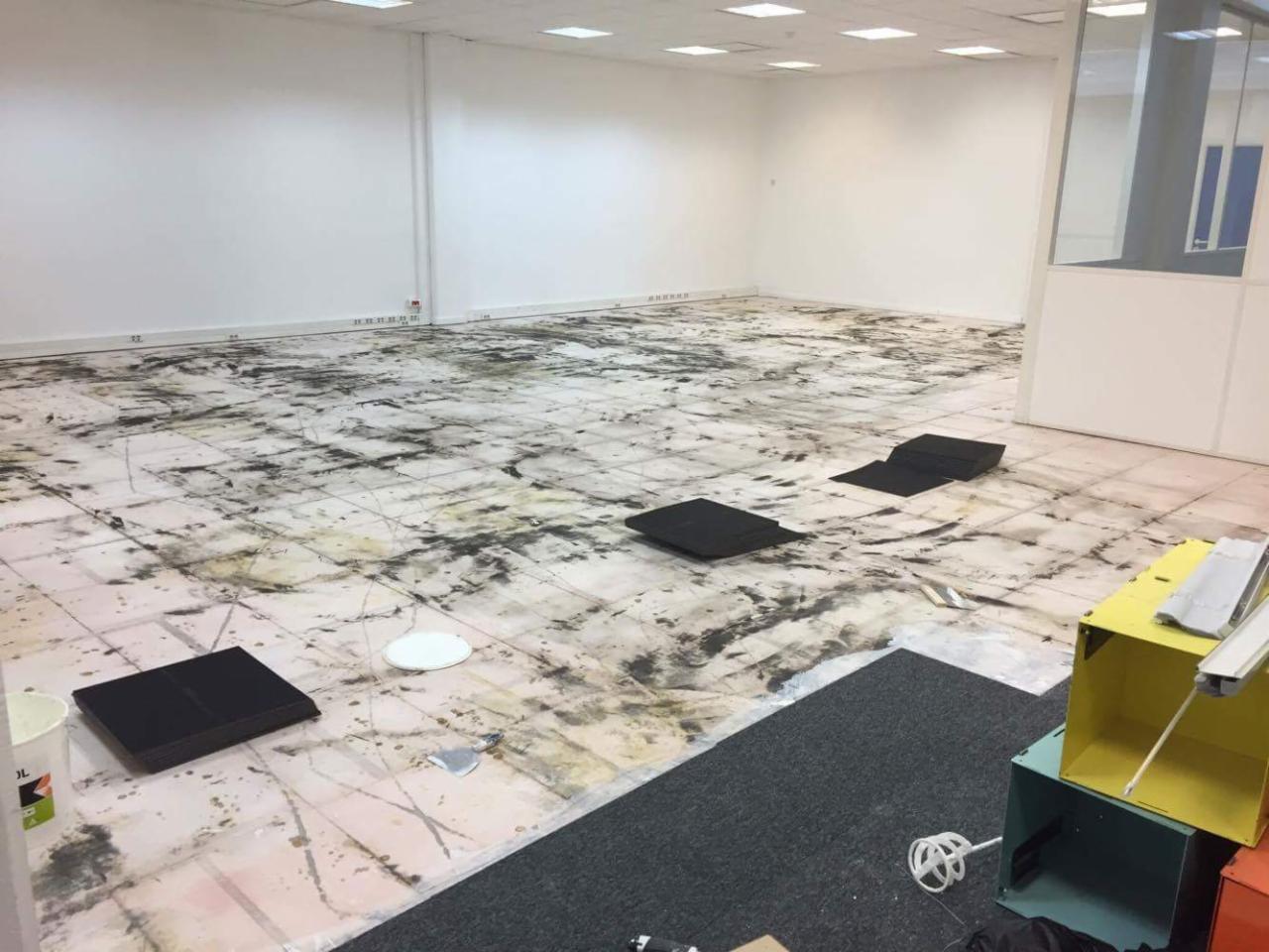 Przygotowanie podłoża w biurze przed pracami wykończeniowymi, folia ochronna na podłodze z widocznymi zabrudzeniami, czarne podkładki, puszki farb i narzędzia malarskie w tle, oświetlenie sufitowe.