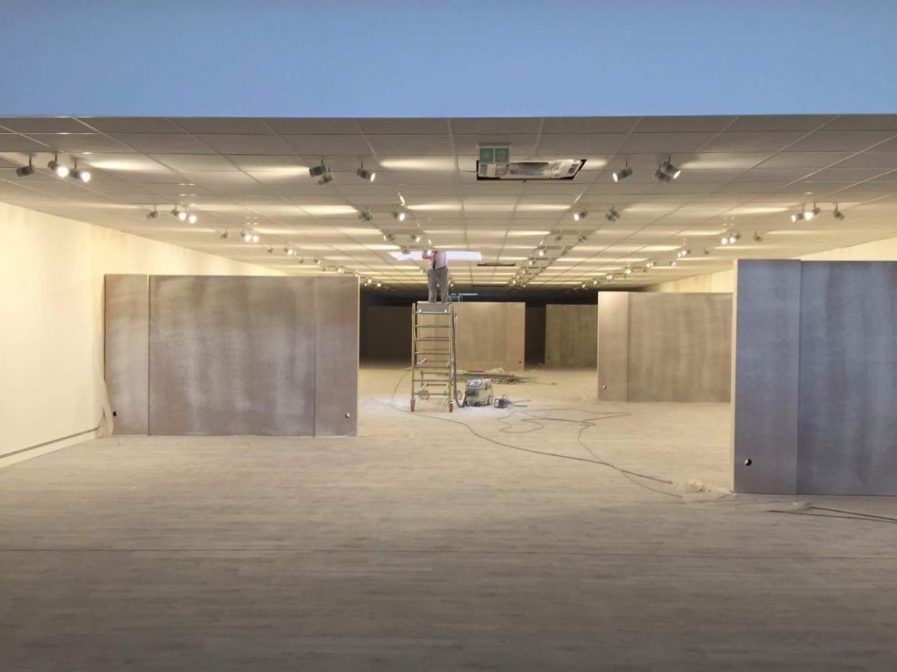 Przestronne wnętrze biurowe w trakcie adaptacji: widoczne płyty ścienne z surowym wykończeniem, drabina z pracownikiem przy suficie, liczne reflektory sufitowe, kabel zasilający na podłodze, ślady...