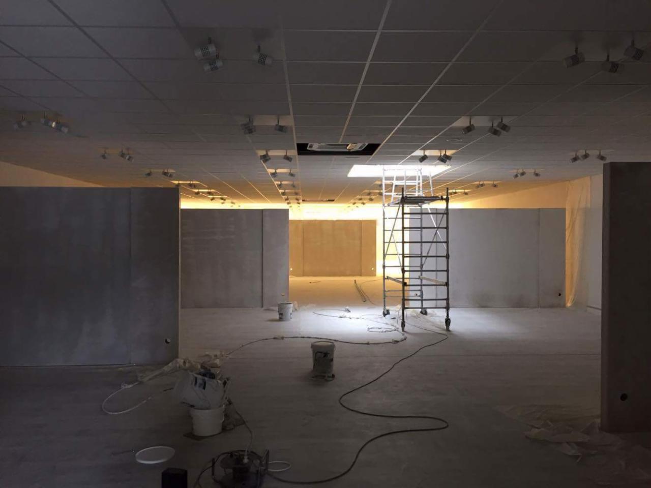 Wnętrze w trakcie prac wykończeniowych: surowy beton na ścianach, metalowy ruszt na środku, widoczne puszki z farbą i przewody, oświetlenie punktowe na suficie rastrowym.