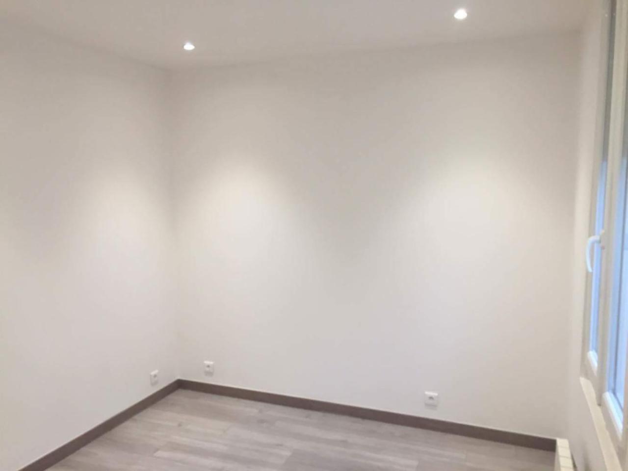 Pomalowane na biało puste pomieszczenie z szarą podłogą, listwami przypodłogowymi i oknem po prawej stronie, oświetlone dwoma punktowymi lampami w suficie.