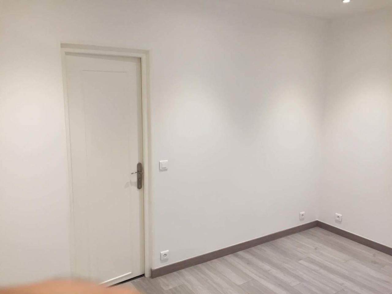 Pusty, świeżo pomalowany pokój z białymi ścianami, drewnopodobną podłogą i białymi drzwiami z brązową klamką, widoczne gniazdka elektryczne przy podłodze.