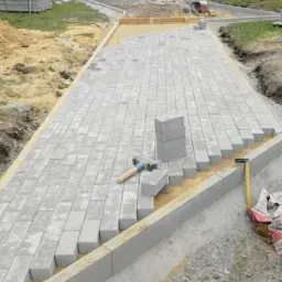 Fragment ścieżki z szarej kostki brukowej w trakcie układania, widoczne narzędzia i stos kostki, obrzeża betonowe, teren przygotowany pod dalsze prace brukarskie.