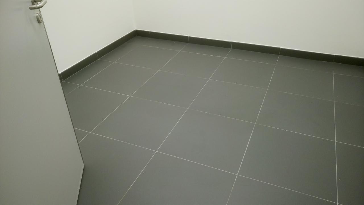 Szare płytki podłogowe z fugami, listwa przypodłogowa i fragment białych ścian oraz drzwi z klamką.