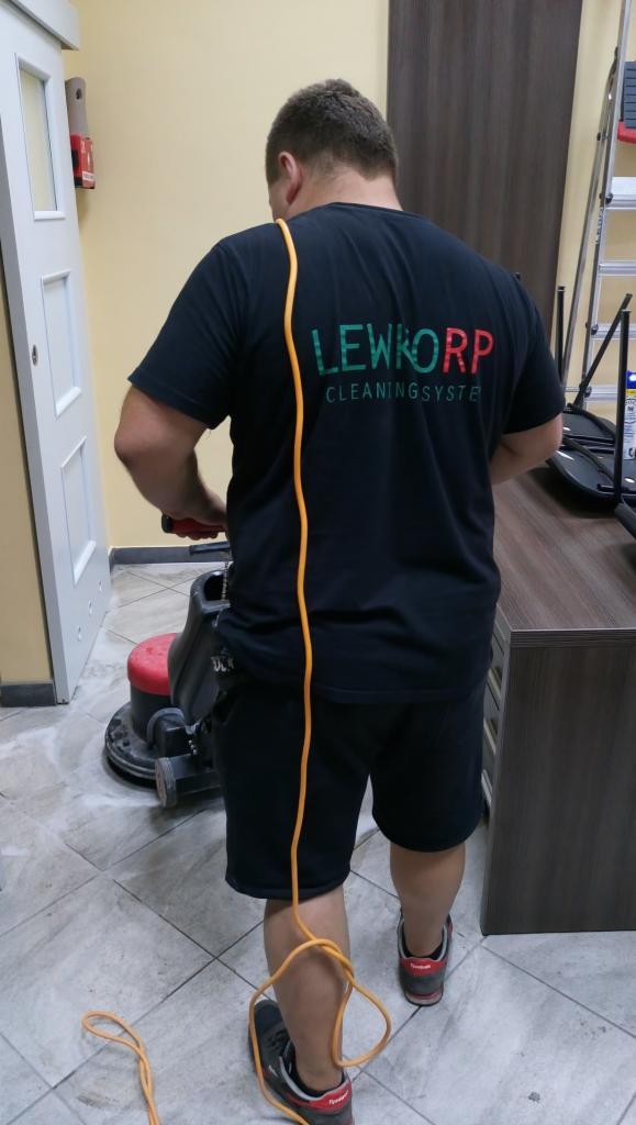 Pracownik w czarnej koszulce z logo LEWKORP Cleaning System obsługuje maszynę do czyszczenia podłóg na jasnych płytkach podłogowych, z widocznym przewodem zasilającym.