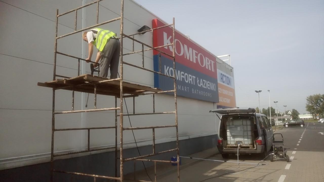 Pracownik w kamizelce na rusztowaniu myje elewację budynku komercyjnego z logo sieci handlowej, używając myjki ciśnieniowej zasilanej z samochodu dostawczego z pojemnikiem na wodę.