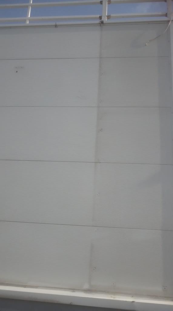 Fragment białej, panelowej ściany z widocznymi poziomymi liniami łączeń, górą widoczna metalowa konstrukcja, prawdopodobnie balustrada.