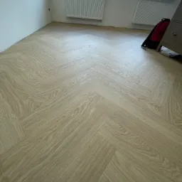 Jasna, dębowa podłoga ułożona w jodełkę francuską w nowym wnętrzu z grzejnikami i plecakiem w rogu.