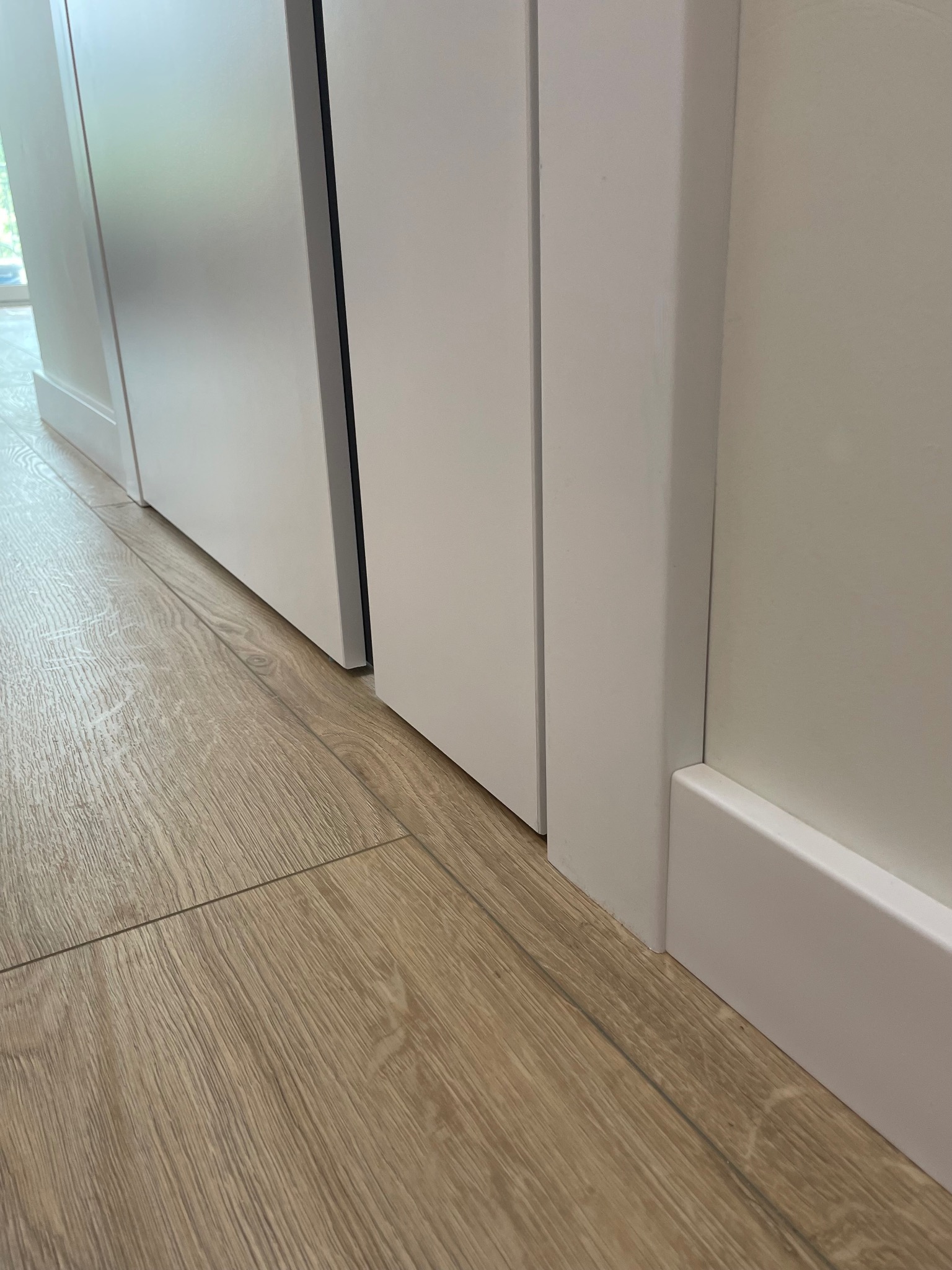 Fragment podłogi z paneli w odcieniach beżu, widoczny przy białej listwie przypodłogowej i framudze drzwi, z delikatnym światłem padającym z okna.