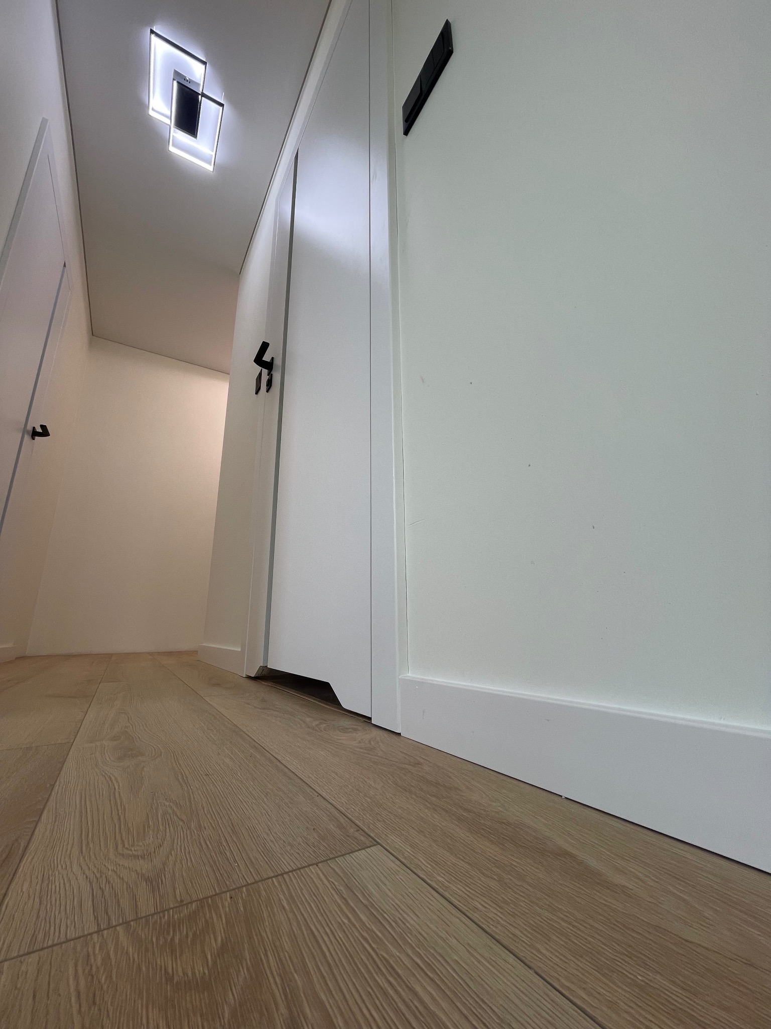 Wnętrze korytarza z panelami podłogowymi w odcieniach dębu, białymi drzwiami z czarnymi klamkami i nowoczesnym oświetleniem sufitowym.