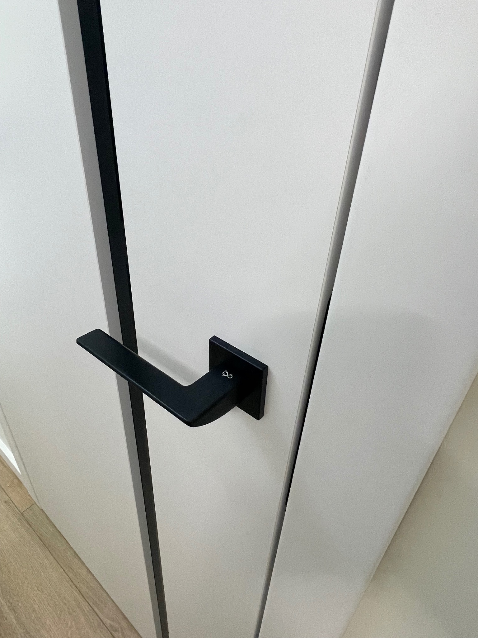 Białe drzwi wewnętrzne z czarną, matową klamką o kwadratowym designie, widoczny fragment drewnianej podłogi.