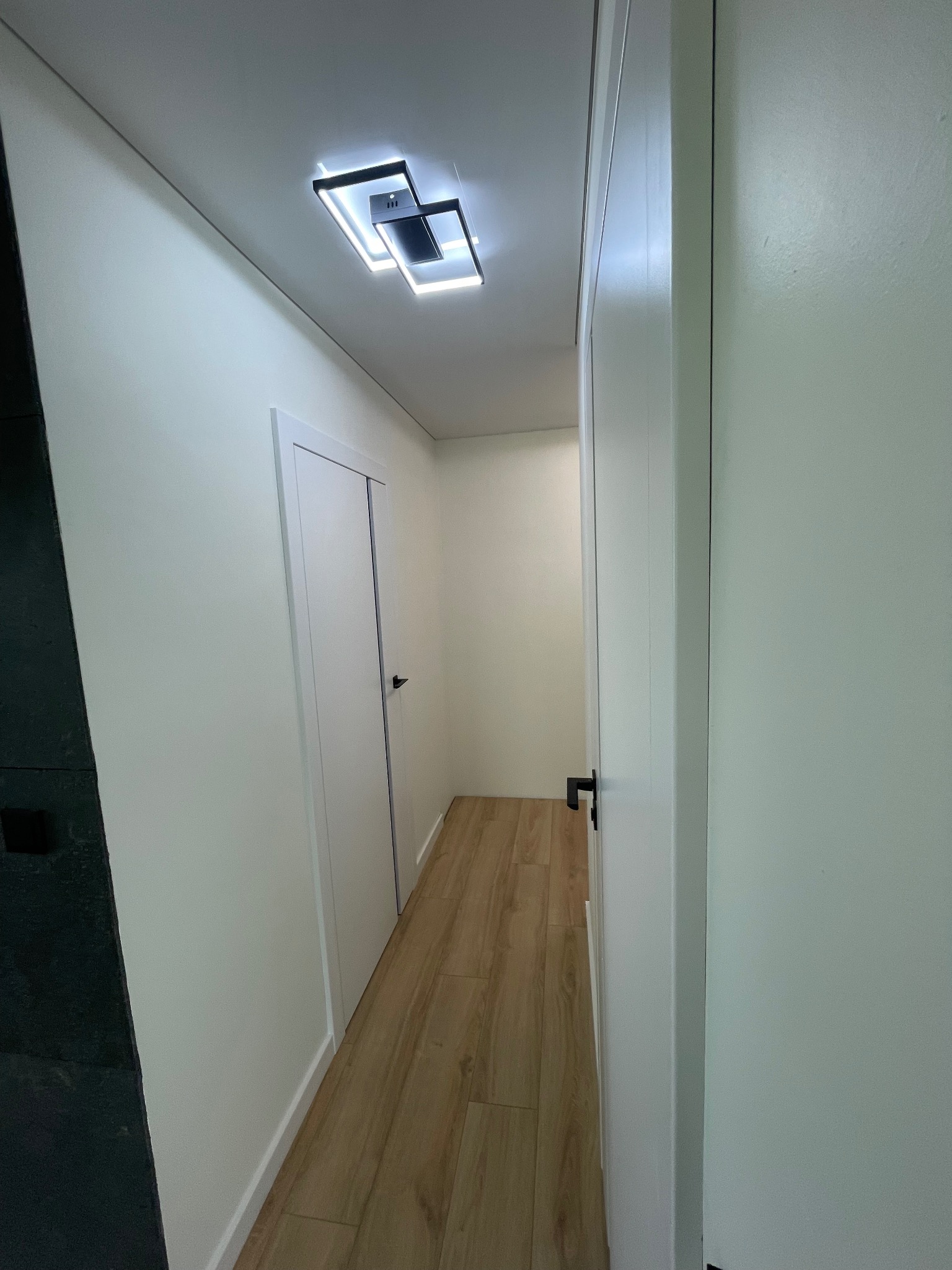 Nowoczesne wnętrze przedpokoju z drewnianą podłogą, białymi ścianami i drzwiami z czarnymi klamkami oraz designerską lampą sufitową.