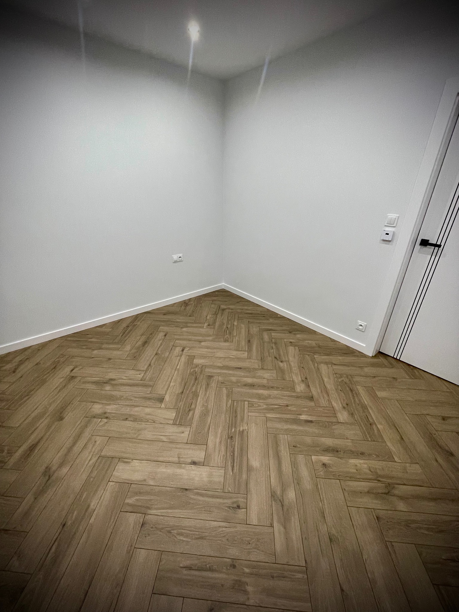 Wnętrze z ułożoną podłogą w jodełkę z paneli imitujących drewno, białe ściany z listwami przypodłogowymi i gniazdkami, białe drzwi z czarną klamką.