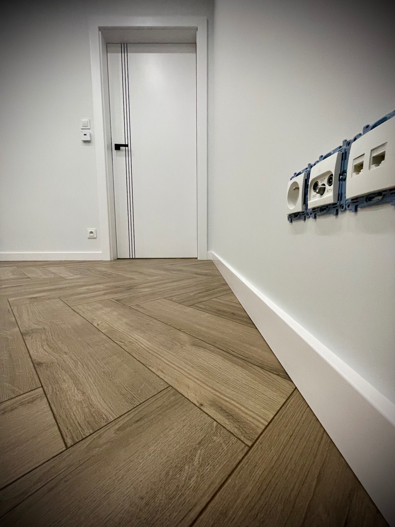 Ułożona w jodełkę podłoga z paneli w odcieniach brązu, biała listwa przypodłogowa, białe drzwi wewnętrzne z czarnymi paskami, gniazdka elektryczne i antenowe w trakcie montażu na białej ścianie.