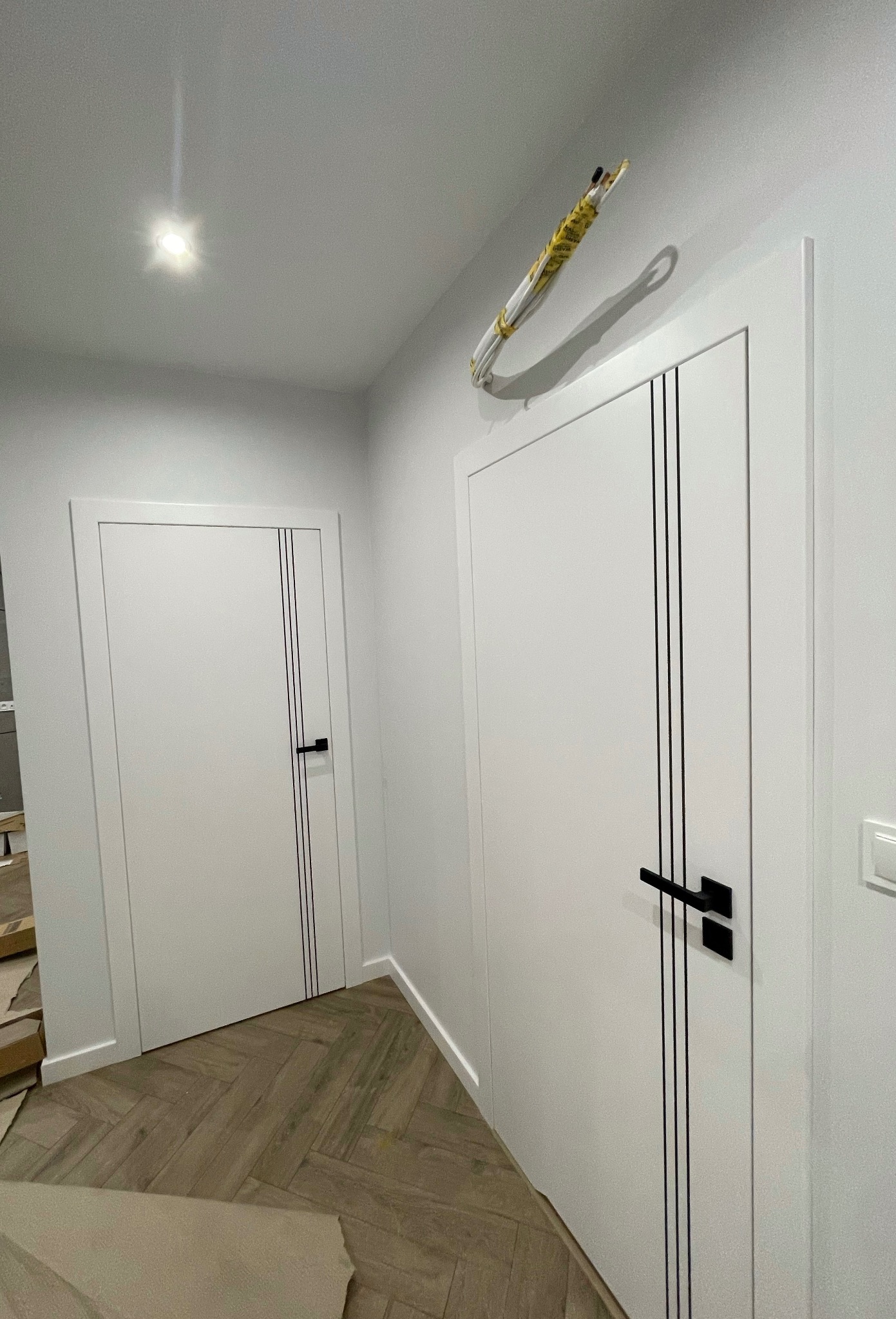Dwa białe skrzydła drzwiowe z czarnymi akcentami w postaci trzech pionowych linii i klamki, widoczne przewody elektryczne zwisające nad jednym z nich, podłoga ułożona w jodełkę.