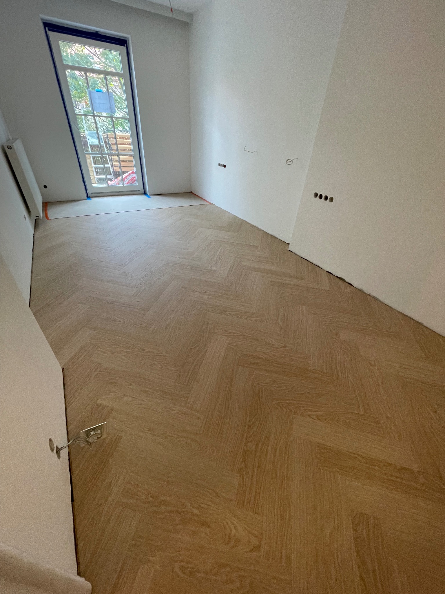 Ułożona podłoga w jodełkę z jasnego drewna, widoczna niezagruntowana ściana i okno z zabezpieczeniem, w trakcie remontu.