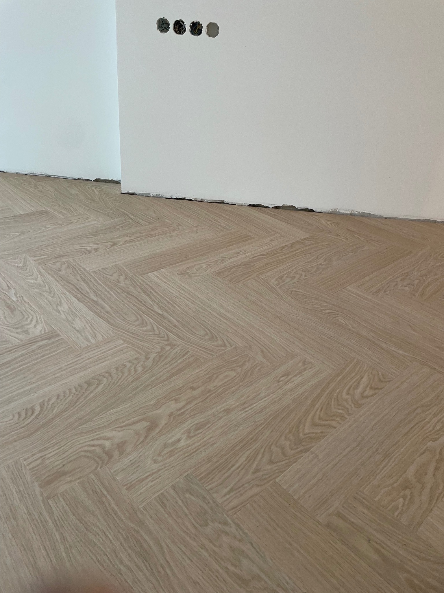 Ułożona podłoga winylowa w jodełkę, odcień dąb rumiany, widoczne otwory na gniazdka elektryczne w ścianie.