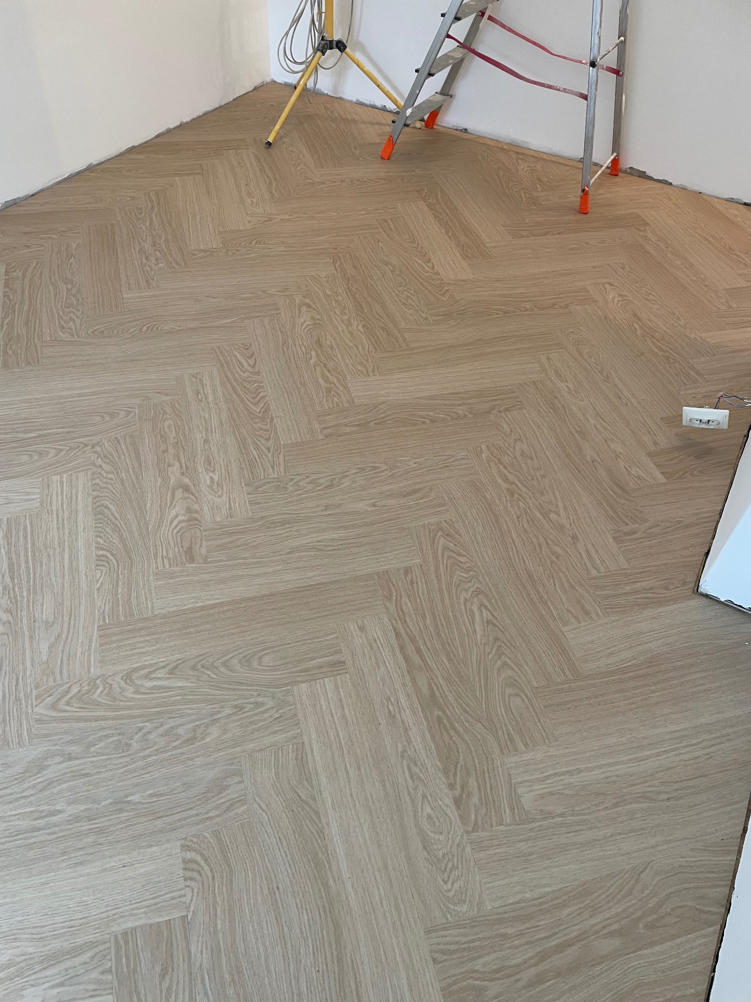 Ułożona podłoga winylowa Quick Step w jodełkę Dąb Rumiany, widoczna lampa robocza i drabina w tle, ślady montażu przy ścianach.