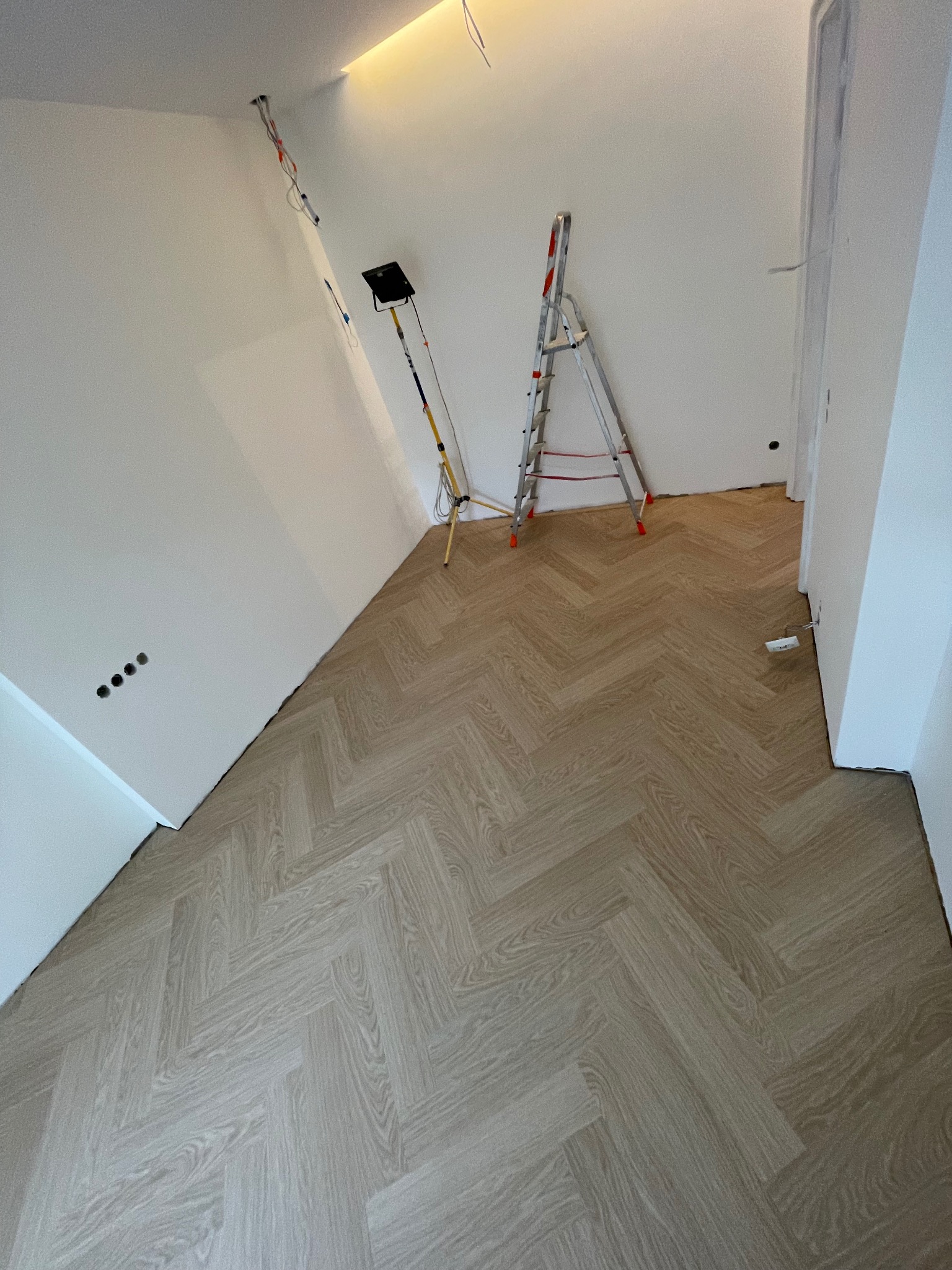 Układanie podłogi winylowej Quick Step w jodełkę dębową rumianą, widoczna drabina i lampa budowlana w trakcie montażu.