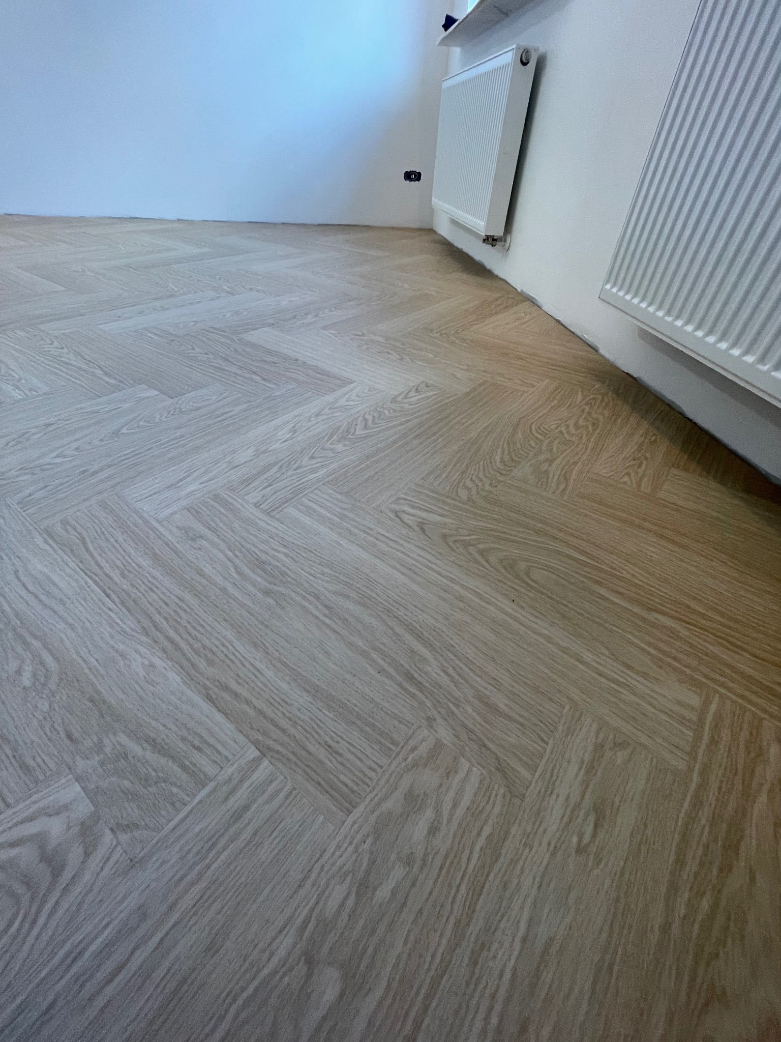 Ułożona podłoga winylowa w jodełkę, odcień dąb rumiany, widoczne grzejniki przy ścianie i fragment białej ściany.