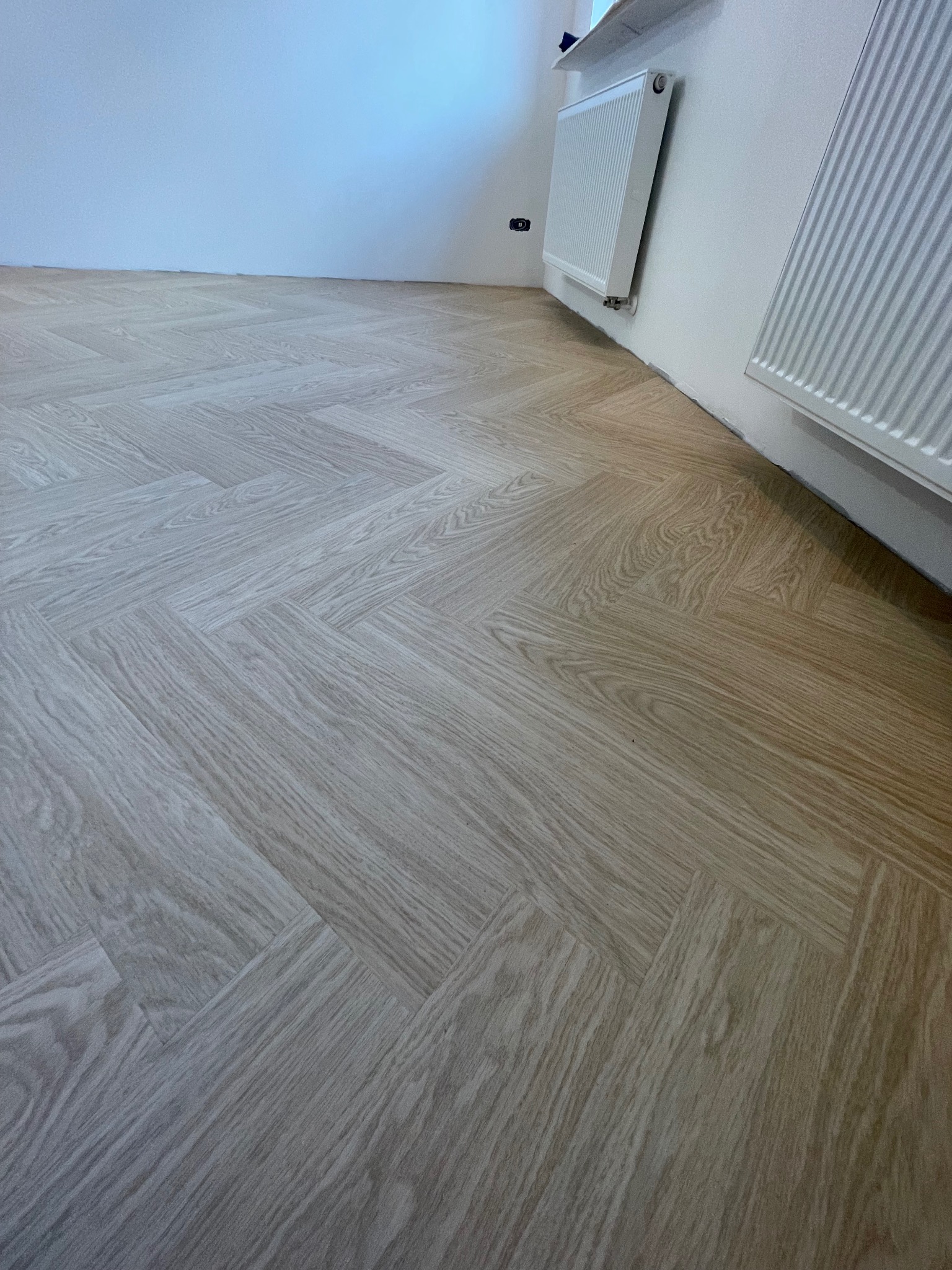 Ułożona podłoga winylowa w jodełkę o jasnym odcieniu dębu rumianego, widoczny fragment ściany z grzejnikiem i gniazdkiem elektrycznym.