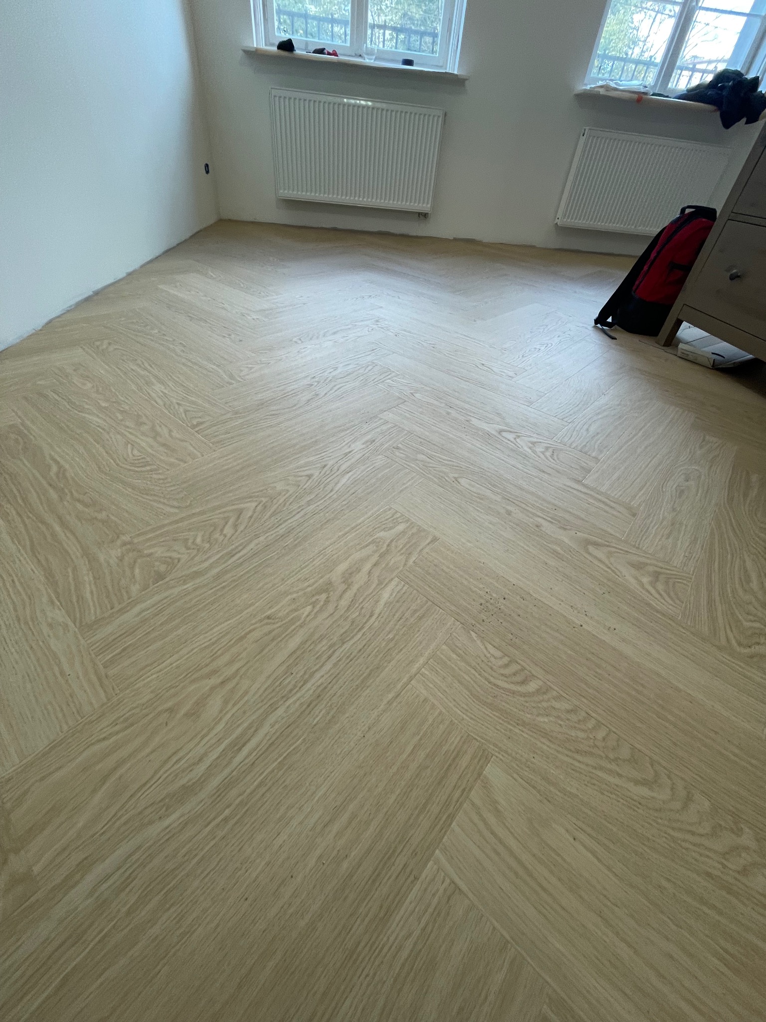 Jasna, dębowa podłoga ułożona w jodełkę francuską w nowym wnętrzu z grzejnikami i plecakiem w rogu.