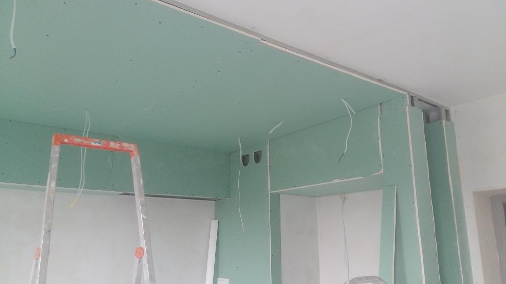 Montaż płyt gipsowo-kartonowych na suficie i ścianach, widoczna metalowa konstrukcja, przewody elektryczne zwisające z sufitu i pomarańczowa drabina.