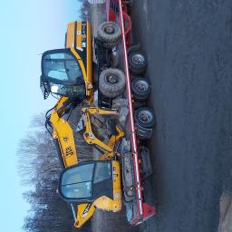 Usługi Transportowe - Dwa żółte mini-koparki JCB załadowane na czerwoną niskopodwoziową przyczepę transportową, widok z boku na tle drzew bez liści i pochmurnego nieba.