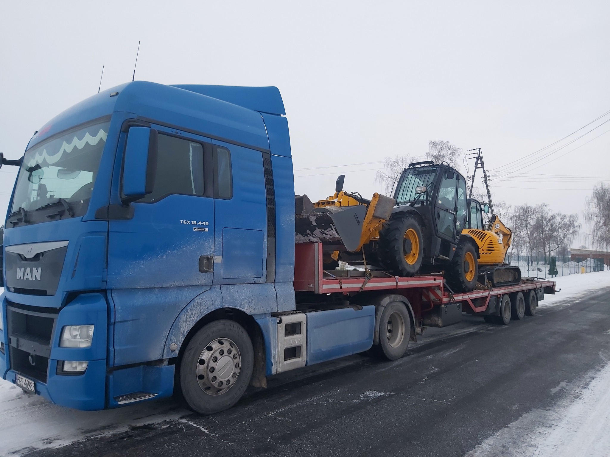 Niebieski ciągnik siodłowy MAN przewozi na niskopodwoziowej platformie żółto-czarną koparko-ładowarkę JCB na tle zimowego krajobrazu.