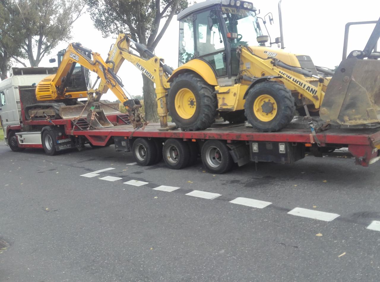 Ciężarówka z naczepą transportuje dwie żółte koparko-ładowarki JCB i New Holland, zabezpieczone pasami, widok z boku na szarej drodze.