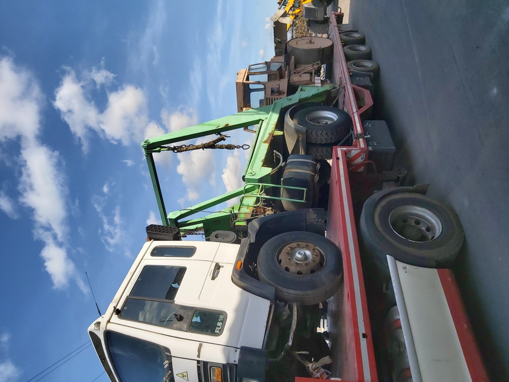 Biała ciężarówka laweta z zielonym dźwigiem przewozi stary, beżowy kombajn na tle błękitnego nieba z chmurami.