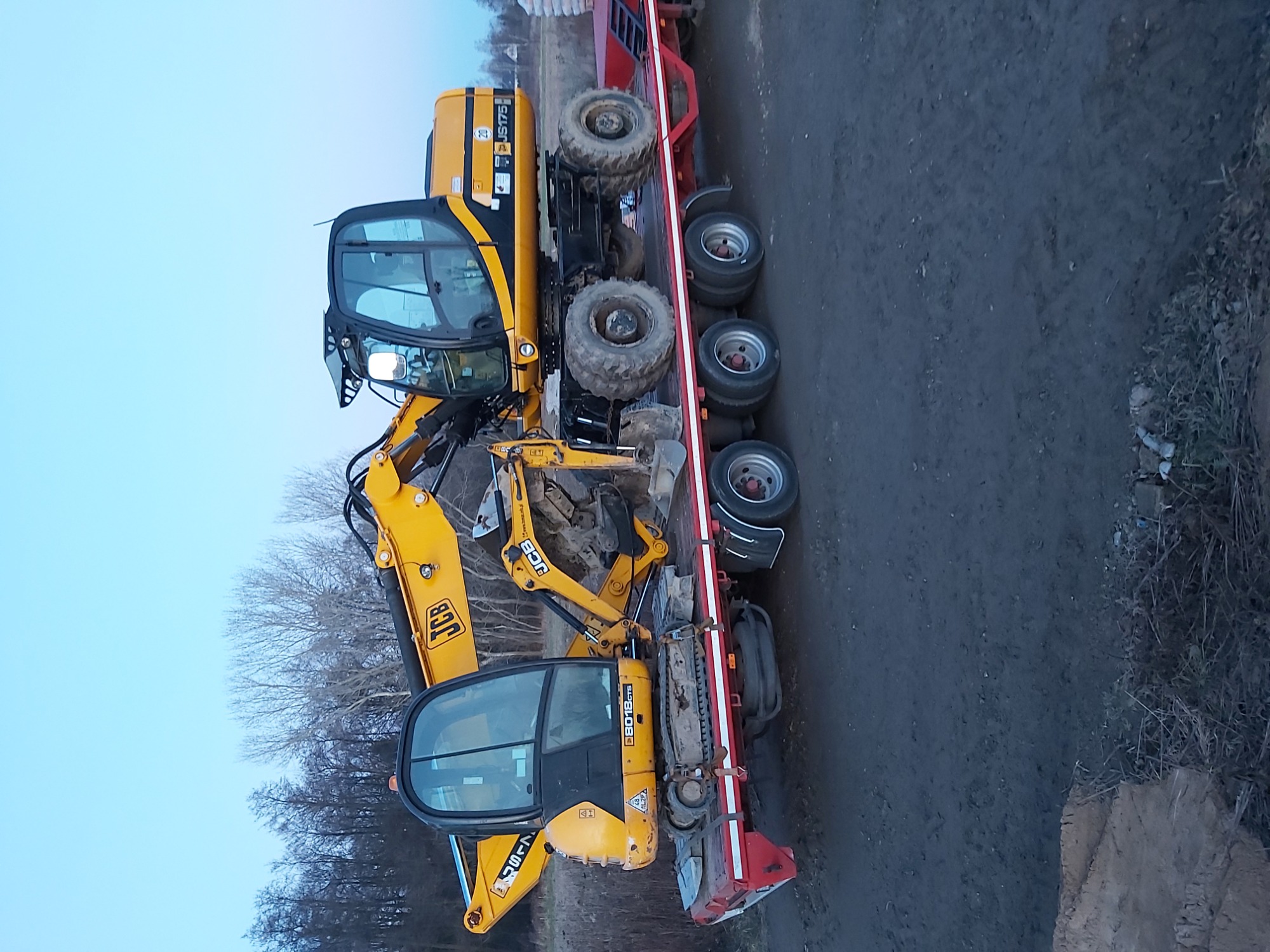 Dwa żółte mini-koparki JCB załadowane na czerwoną niskopodwoziową przyczepę transportową, widok z boku na tle drzew bez liści i pochmurnego nieba.