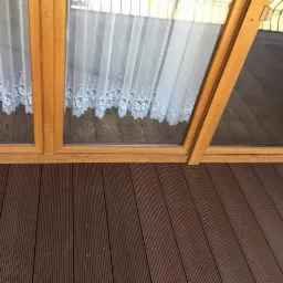 Brunatna deska kompozytowa na balkonie, widok od dołu okna z koronkową firanką.
