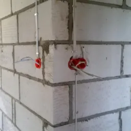 Surowa ściana z betonu komórkowego z zamontowanymi puszkami elektrycznymi w kolorze czerwonym i wystającymi przewodami, widoczne metalowe rurki instalacyjne mocowane do ściany klamrami.