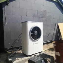 Biała pompa ciepła Bosch zamontowana na betonowej podstawie przy budynku ocieplonym szarym styropianem, widoczne elementy montażowe i beczka.