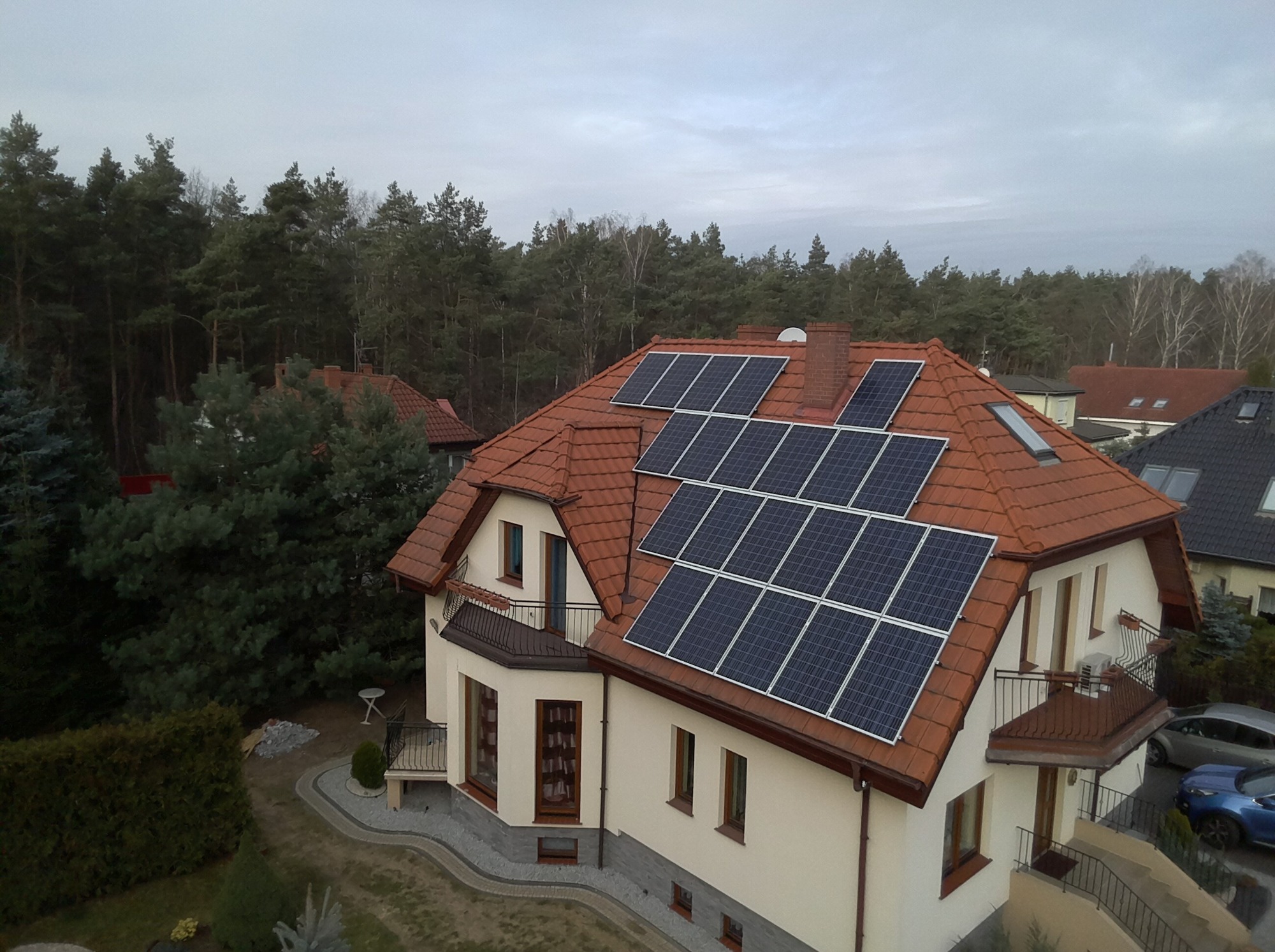 Dom jednorodzinny z czerwoną dachówką, na której zamontowano panele fotowoltaiczne; widok z góry, w tle las.