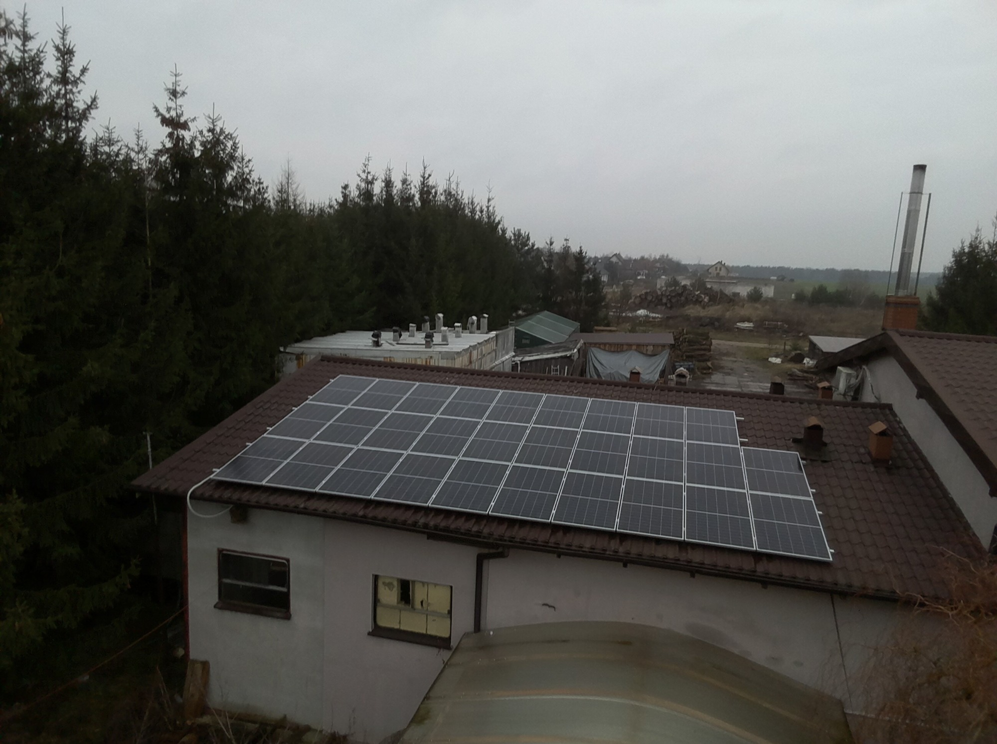 Panel fotowoltaiczny na dachu budynku gospodarczego z brązową dachówką, widok z góry w pochmurny dzień, w tle las iglasty i zabudowania.