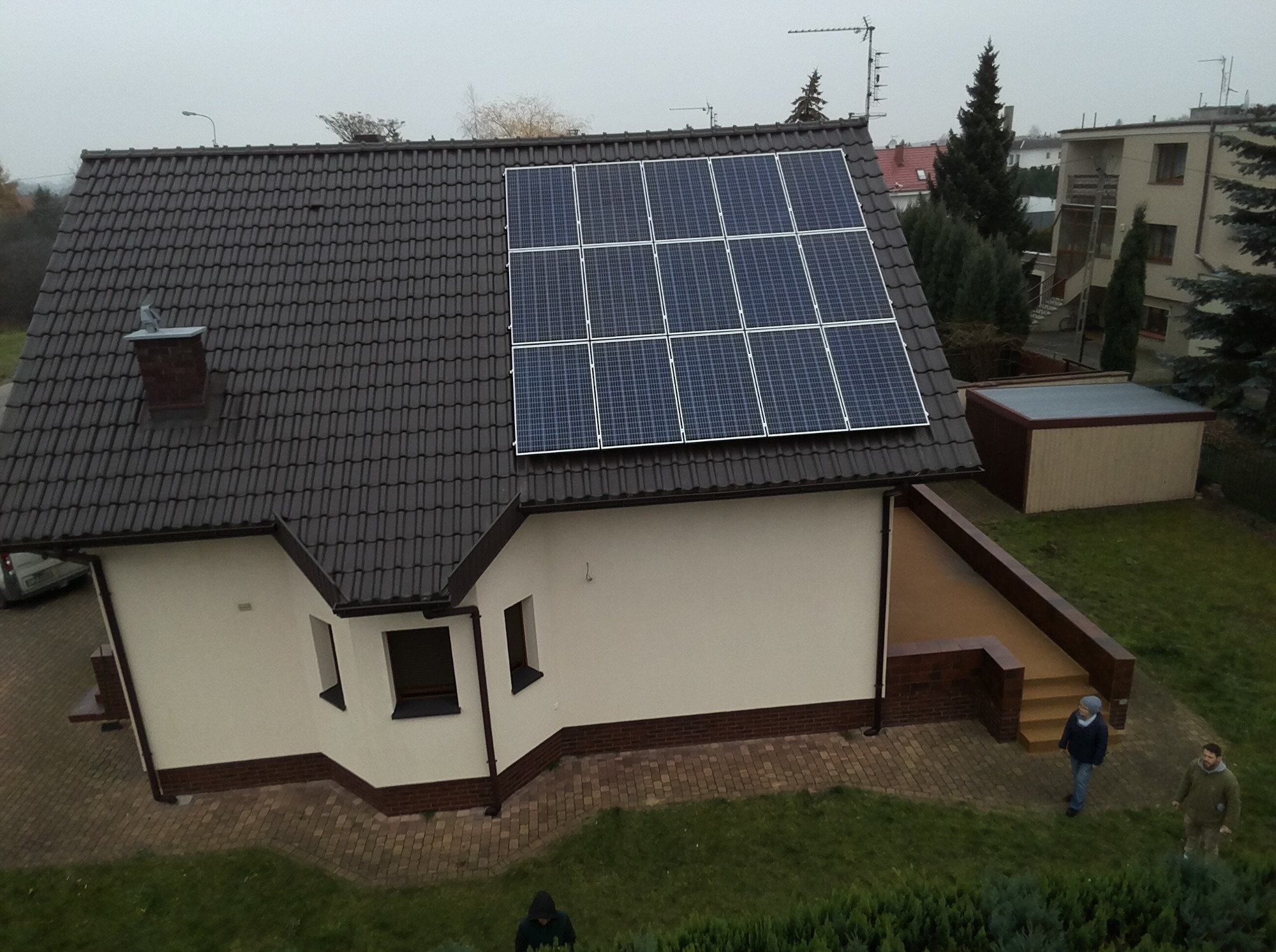 Widok z góry na dom z ciemnym dachem, na którym zainstalowano panele słoneczne; na trawniku widać dwie osoby.