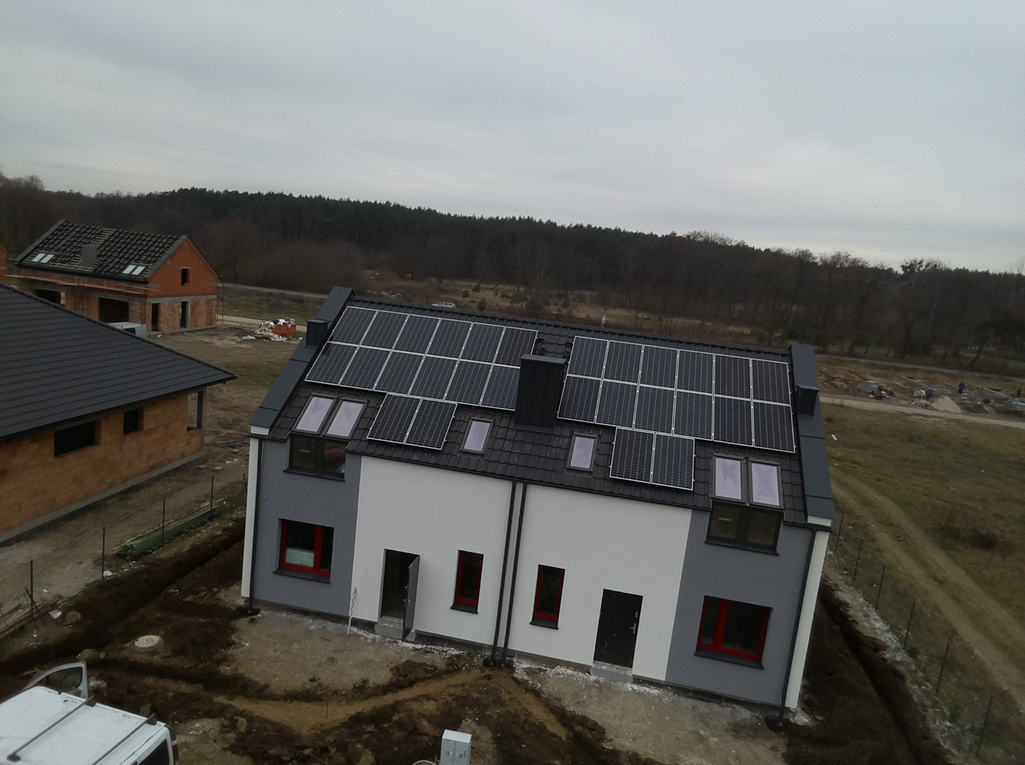 Nowoczesny dom dwurodzinny z panelami fotowoltaicznymi na dachu, widok z góry na tle lasu i pochmurnego nieba.