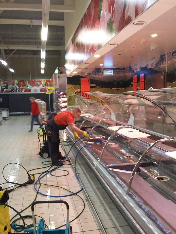 Pracownik w pomarańczowej koszulce i żółtych rękawicach myje ciśnieniowo witrynę chłodniczą w supermarkecie, z widocznymi przewodami i urządzeniem wysokociśnieniowym na podłodze z płytek.