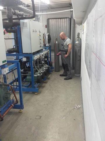 Mężczyzna w kombinezonie roboczym sprawdza instalację chłodniczą z użyciem czerwonego miernika, obok stoją butle z gazem, widok z boku.